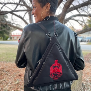 May include: Black triangular sling bag with a red embroidered crest. The bag has a zipper closure and adjustable strap. The crest features Greek letters and a shield design. The bag is worn over the shoulder.