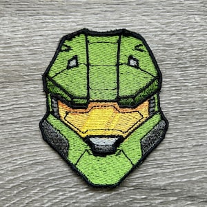 Può includere: Toppa ricamata raffigurante un casco verde e giallo con contorni neri. La toppa ha un aspetto dettagliato e strutturato ed è probabilmente destinata all'applicazione su abbigliamento o accessori. Il design ricorda un tema fantascientifico o videoludico.