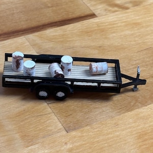 Puede incluir: Un remolque negro en miniatura con cuatro pequeños barriles cilíndricos. Los barriles son blancos con marcas marrones y rojas. El remolque tiene una plataforma de madera, sobre un fondo de madera.