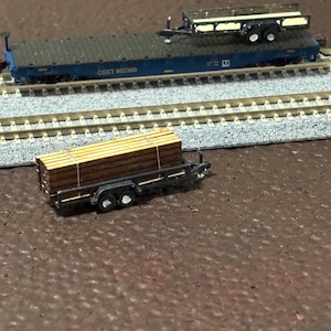 May include: A model train flatcar with the text CSXT 602360, featuring a small empty trailer on its deck. In the foreground, a separate small trailer carries a stack of brown wooden planks secured with white straps, all set on model train tracks.