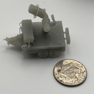 May include: A small, unpainted gray resin 3D-printed model of a trailer with a mounted cannon-like apparatus, positioned next to a standard United States dime for scale comparison, showing the miniature size of the detailed plastic object.