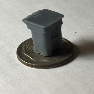 May include: A miniature gray trash bin with two small wheels, resting on a quarter coin. The small trash can has a rectangular shape with a hinged lid. The coin is silver in color.