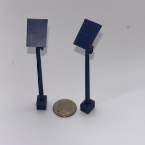 May include: Two miniature dark blue solar panel models, each featuring a rectangular panel mounted on a thin pole with a square base, positioned on either side of a small circular silver coin for scale comparison.
