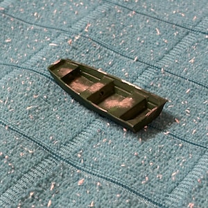 Puede incluir: Un pequeño barco de juguete verde con fondo plano y dos asientos. El barco está hecho de un material sólido y tiene un diseño sencillo. Descansa sobre una superficie azul texturizada.