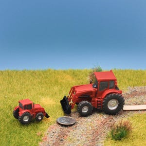 May include: Two red toy tractors are set on a grassy surface against a blue sky. The larger tractor features a front loader and large tyres. A small coin is placed near the tractors. The scene suggests a miniature farm environment.