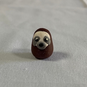 May include: A small brown sloth figurine with a white face and black eyes. The sloth is made of clay and has a rounded shape.