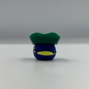 May include: A small blue bird figurine with a yellow beak and black eyes, wearing a green hat.