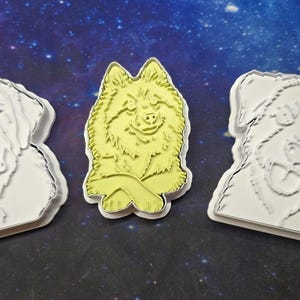 Op de afbeelding: Drie koekjesvormpjes in de vorm van honden. Twee zijn wit en tonen gedetailleerde contouren van hondengezichten. De middelste vorm is lichtgeel en toont een hond met gekruiste voorpoten. Elke vorm heeft een opstaande rand voor het snijden.