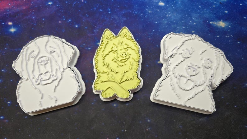 Op de afbeelding: Drie koekjesvormpjes in de vorm van honden. Twee zijn wit en tonen gedetailleerde contouren van hondengezichten. De middelste vorm is lichtgeel en toont een hond met gekruiste voorpoten. Elke vorm heeft een opstaande rand voor het snijden.