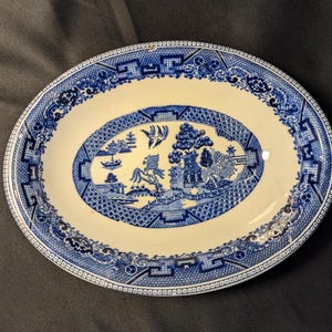 Op de afbeelding: Een ovale blauwe en witte keramische serveerschaal met een wilgenmotief. De schaal heeft een witte achtergrond met een blauwe rand en een blauw wilgenboomontwerp in het midden.