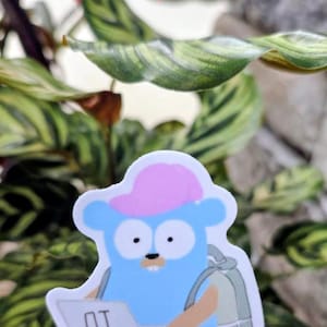 May include: A blue sticker featuring a cartoon character with a pink hat and backpack, holding a laptop with "AI" on the screen. A coffee cup is next to the laptop. The sticker is set against a backdrop of green and red leaves.