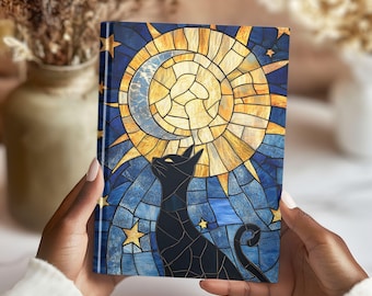 May include: A hardcover journal featuring a stained-glass design of a black cat gazing at a yellow sun and stars against a blue background. The journal's spine is dark blue. The cover is held by hands.