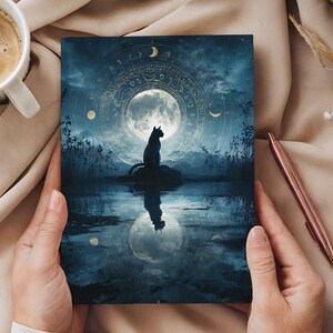 May include: A dark blue art print featuring a black cat silhouette perched on a rock, reflecting in the water. The background includes a full moon, celestial symbols, and a night sky. The print is held by hands.