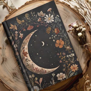 May include: A hardcover journal with a dark cover featuring a crescent moon and floral design. The moon is surrounded by stars and flowers in shades of orange, white, and brown. The journal is resting on a wooden surface.