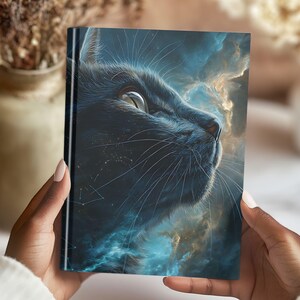 May include: A hardback journal with a detailed illustration of a black cat's face against a backdrop of swirling blue and gold clouds. The cat's eye is bright, and whiskers are visible. The journal is held in hands.