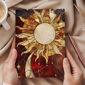 May include: A stained glass style notebook cover with a large sun design in shades of gold, orange, and red. The sun has a white center and is surrounded by a dark red background with gold stars.