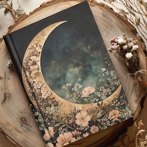 May include: A black journal with a crescent moon design. The moon is filled with a dark blue and green sky and surrounded by floral illustrations in shades of pink, white, and beige. The journal is resting on a wooden surface.