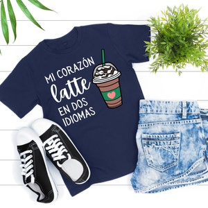 May include: Navy blue t-shirt with the text "MI CORAZÓN latte EN DOS IDIOMAS" and a graphic of a coffee drink. The shirt is paired with denim shorts and black and white sneakers.