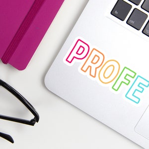 May include: A laptop with a sticker that says "PROFE" in colorful outlined letters. A magenta notebook and black glasses are also on the white surface.