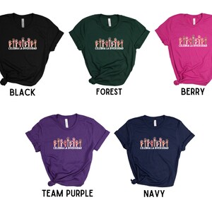 May include: A collection of t-shirts in various colors: black, forest green, berry pink, team purple, and navy blue. Each shirt features a graphic of raised hands in different skin tones above the text "CELEBRA LA DIVERSIDAD".