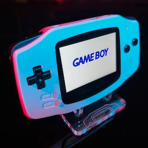 Puede incluir: Una consola de videojuegos portátil Game Boy Advance azul y blanca con un soporte de plástico transparente. La pantalla muestra las palabras "GAME BOY" en texto azul.