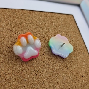 May include: Two colorful push pins in the shape of a paw print and a cloud. The paw print is orange, yellow, pink, and white. The cloud is rainbow colored.