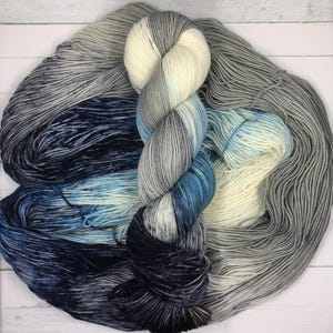 May include: A skein of hand-dyed yarn in a gradient of colors. The yarn transitions from dark navy blue to light blue, gray, and cream. The yarn is soft and appears to be made of natural fibers, perfect for knitting or crocheting projects.
