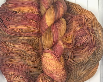 May include: A skein of hand-dyed yarn in shades of orange, red, and brown. The yarn is arranged in a twisted skein and a loose pile, showcasing the color variations. The colors blend together, creating a warm, autumnal feel, perfect for knitting or crocheting projects.