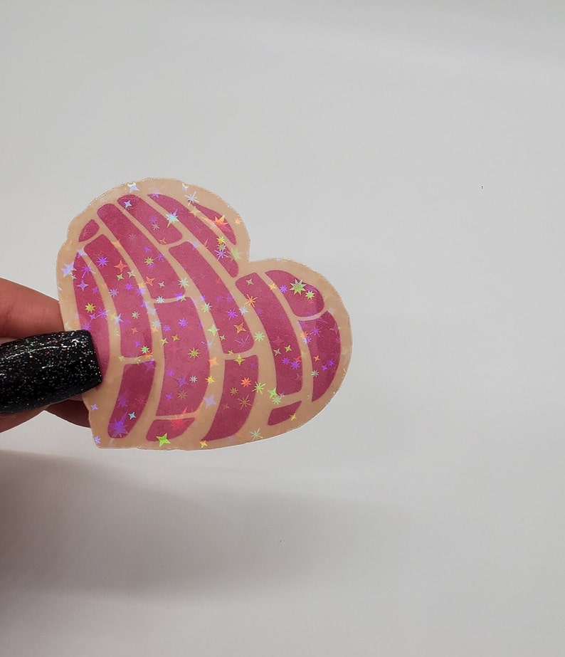 Pu&ograve; includere: Un adesivo rosa a forma di cuore con un motivo di linee e stelle. L'adesivo ha una finitura olografica che brilla di diversi colori.