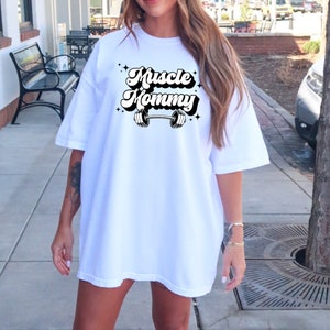 May include: White oversized t-shirt with a black graphic that says "Muscle Mommy" with a barbell underneath the text.