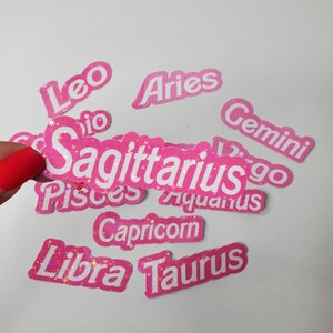 May include: Pink and white glitter stickers with the names of the zodiac signs: Leo, Aries, Gemini, Sagittarius, Pisces, Aquarius, Capricorn, Libra, and Taurus.