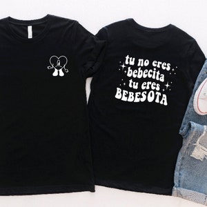 May include: Two black t-shirts with white text and graphics. The front shirt has a cartoon heart with arms and legs. The back shirt has the text "tu no eres bebecita tu eres BEBESOTA" with stars on either side.