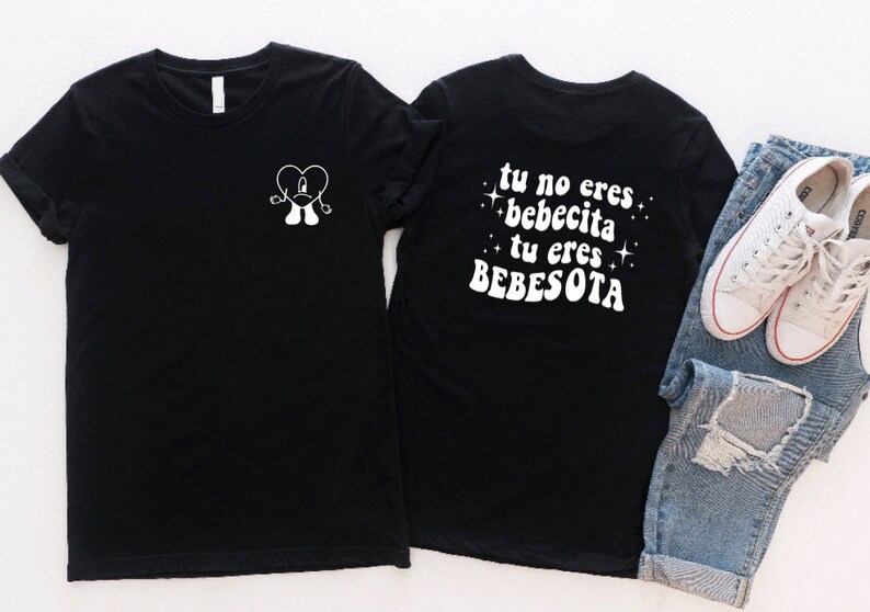 May include: Two black t-shirts with white text and graphics. The front shirt has a cartoon heart with arms and legs. The back shirt has the text "tu no eres bebecita tu eres BEBESOTA" with stars on either side.