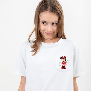 May include: A white t-shirt with a red and black Minnie Mouse graphic on the pocket. The girl wearing the shirt is looking to the side and smiling.