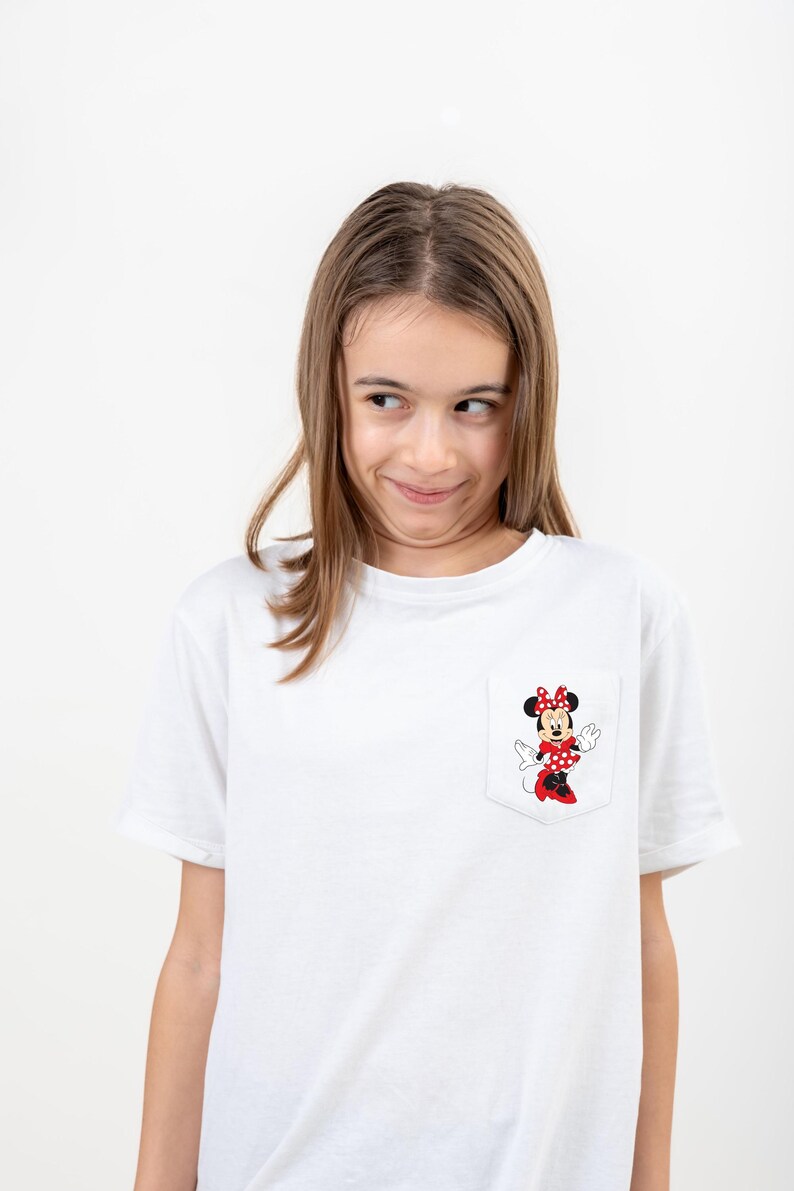 May include: A white t-shirt with a red and black Minnie Mouse graphic on the pocket. The girl wearing the shirt is looking to the side and smiling.