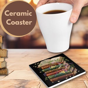 Puede incluir: Un posavasos cuadrado de cerámica con un diseño de pila de libros se encuentra sobre una superficie de piedra. Una taza de café blanca se sostiene sobre el posavasos. Una computadora portátil y una pila de libros también están sobre la mesa. El posavasos tiene las palabras "Ceramic Coaster".