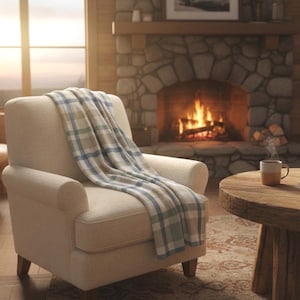 May include: An off-white upholstered armchair featuring a plaid throw blanket with blue, green, and beige stripes draped over the back and seat. The chair has a rounded backrest, padded armrests, and short wooden legs, providing a neutral seating option.