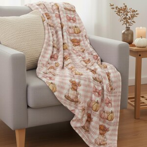 May include: A pink and white plaid throw blanket draped over a gray armchair. The blanket features a repeating pattern of pumpkins, bows, and cow illustrations. A beige pillow rests on the chair. The scene evokes a cozy, autumnal theme.