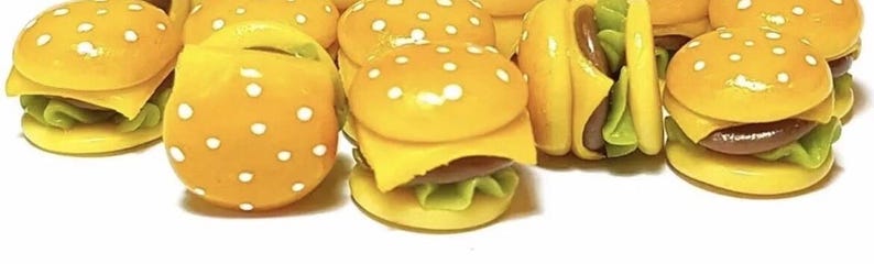 May include: A group of miniature yellow and brown toy hamburgers with white sesame seeds on top. The hamburgers are made of polymer clay and are about the size of a dime.