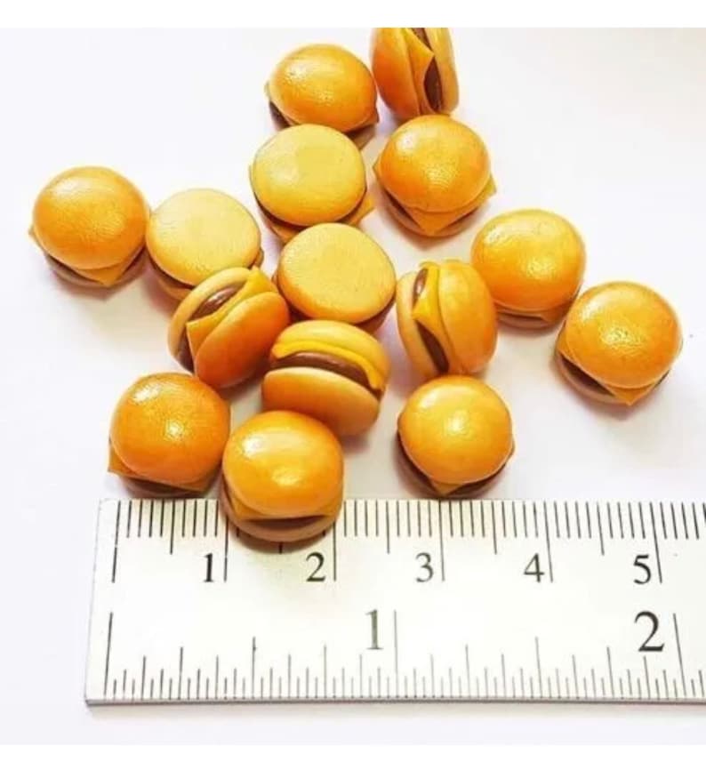 May include: A collection of 12 miniature yellow and brown cheeseburger-shaped charms, measuring approximately 1 inch in diameter, scattered on a white surface next to a ruler.