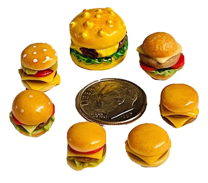 May include: A collection of seven miniature toy hamburgers, each with a sesame seed bun, cheese, lettuce, and tomato. The hamburgers are arranged on a white background.