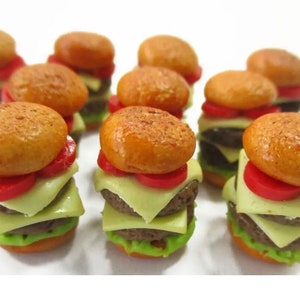May include: Nine miniature toy cheeseburgers with red tomato slices, cheese, and lettuce on a white surface.
