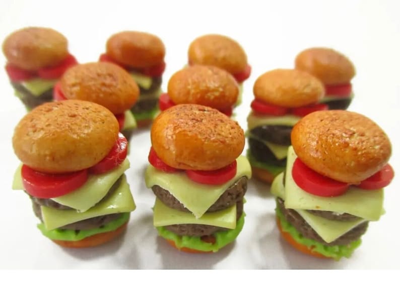 May include: Nine miniature toy cheeseburgers with red tomato slices, cheese, and lettuce on a white surface.