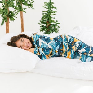 Puede incluir: Una persona joven con un pijama azul y amarillo con estampado de montaña yace en una cama blanca con una almohada blanca. La persona sonríe y mira a la cámara.