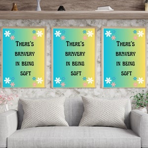 Puede incluir: Tres impresiones enmarcadas con un fondo degradado azul y amarillo y flores blancas. El texto en las impresiones dice "There's bravery in being soft."