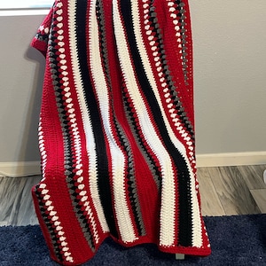May include: A striped crocheted blanket in red, white, black, and gray. The blanket is draped over a chair.