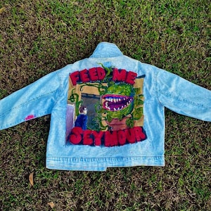 Könnte beinhalten: Hellblaue Jeansjacke mit handgemaltem Design. Das Design zeigt eine große, grüne, fleischfressende Pflanze und die Worte "FEED ME SEYMOUR" in Rot. Die Jacke hat einen klassischen Kragen und eine Knopfleiste.