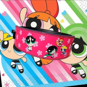 Peut inclure: Ceinture de musculation rose avec une boucle noire et un imprim&eacute; des personnages de Powerpuff Girls. La ceinture a un fond rose avec des fleurs blanches et les personnages sont dans leurs poses caract&eacute;ristiques.