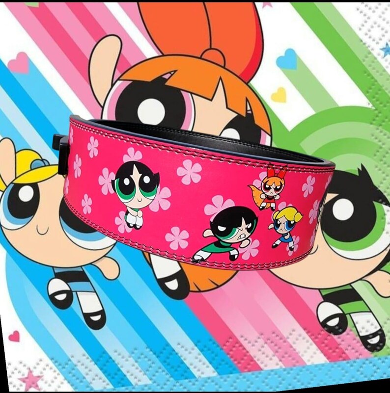 Peut inclure: Ceinture de musculation rose avec une boucle noire et un imprim&eacute; des personnages de Powerpuff Girls. La ceinture a un fond rose avec des fleurs blanches et les personnages sont dans leurs poses caract&eacute;ristiques.