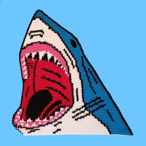 Peut inclure: Une tête de requin en crochet avec la bouche ouverte, réalisée en fil blanc, bleu, rose et noir. Les dents du requin sont visibles et la bouche est grande ouverte.